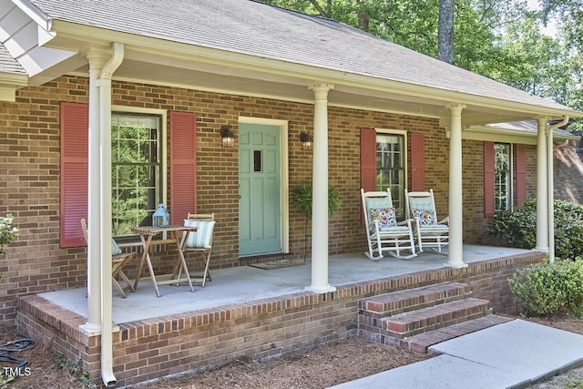 entrance to property featuring brick siding, roof with shingles, and covered porch