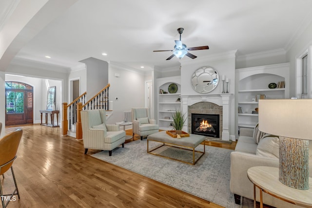 living room featuring a ceiling fan, built in features, a tile fireplace, wood finished floors, and ornamental molding