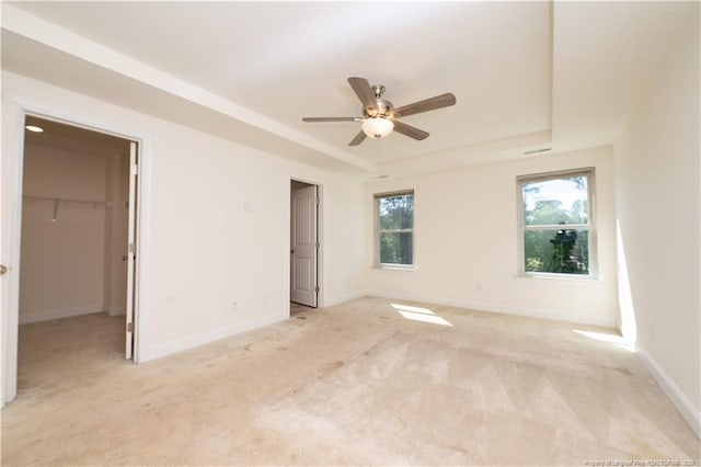 unfurnished bedroom featuring light carpet, baseboards, a walk in closet, ceiling fan, and a raised ceiling