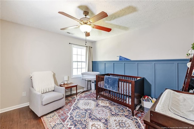 bedroom with a nursery area, ceiling fan, wainscoting, wood finished floors, and visible vents
