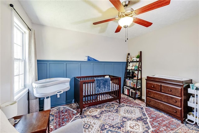 bedroom featuring a crib, multiple windows, a wainscoted wall, and a ceiling fan