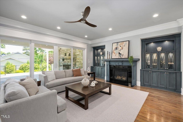 living room featuring crown molding, a ceiling fan, a fireplace with flush hearth, light wood-style flooring, and recessed lighting