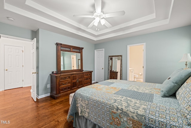 bedroom with wood finished floors, a tray ceiling, ensuite bathroom, baseboards, and ceiling fan