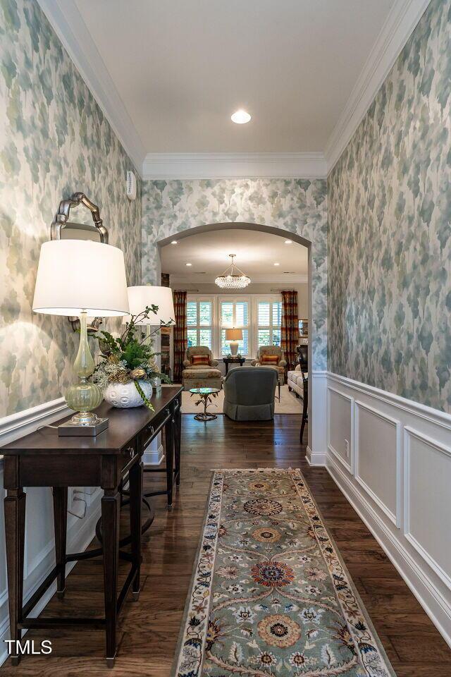 hallway with wallpapered walls, arched walkways, crown molding, dark wood-type flooring, and wainscoting