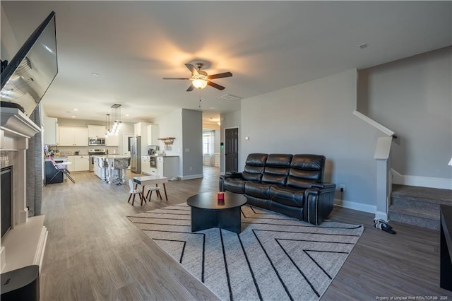 living room featuring a ceiling fan, baseboards, a fireplace with raised hearth, light wood-style floors, and stairs