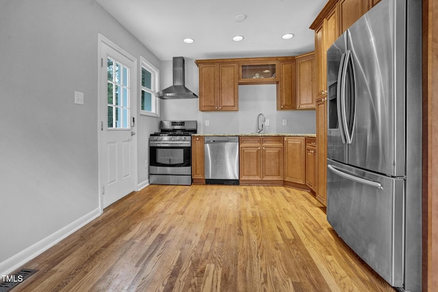 kitchen featuring wall chimney range hood, appliances with stainless steel finishes, brown cabinetry, and light wood-type flooring