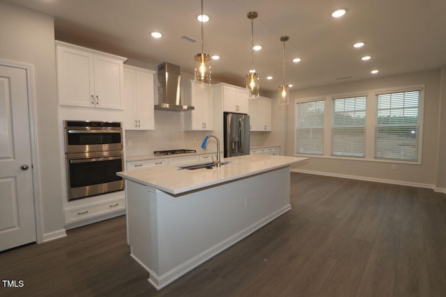 kitchen with wall chimney range hood, a sink, stainless steel appliances, dark wood-type flooring, and a center island with sink