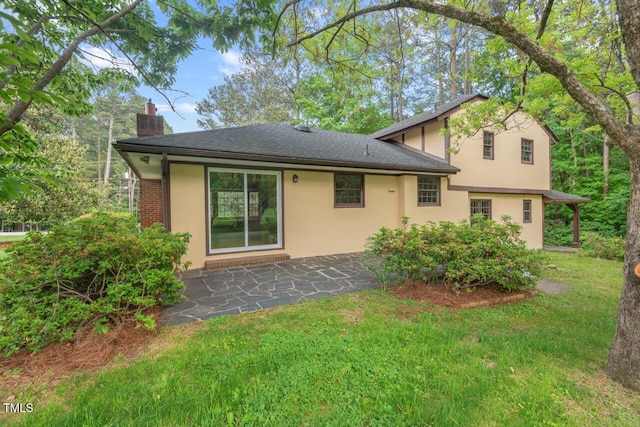 rear view of house with roof with shingles, stucco siding, a chimney, a lawn, and a patio