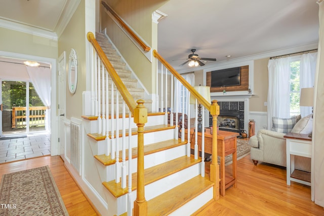 stairs with ornamental molding, ceiling fan, a tile fireplace, wood finished floors, and a wainscoted wall