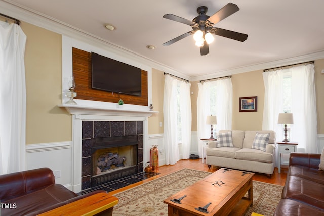 living area featuring a ceiling fan, a tile fireplace, ornamental molding, wood finished floors, and a wainscoted wall