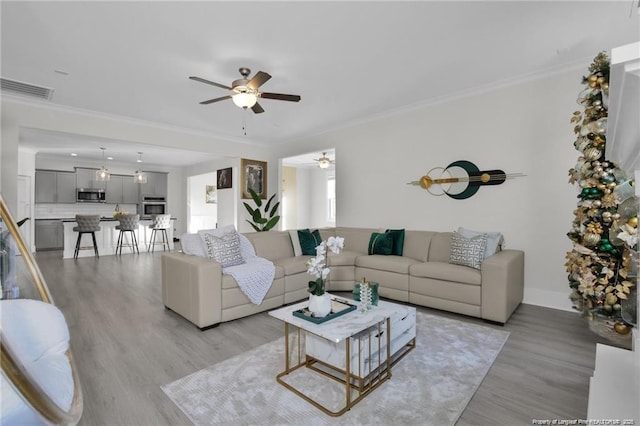 living room with ceiling fan, visible vents, baseboards, ornamental molding, and light wood-style flooring