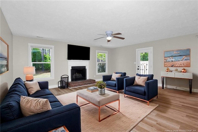 living area with baseboards, ceiling fan, a brick fireplace, and light wood-type flooring