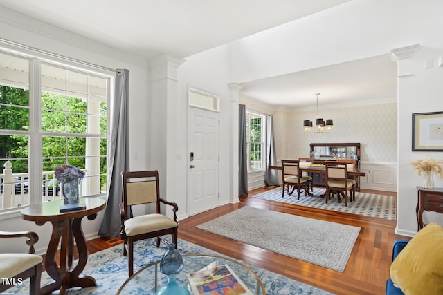 living room with wallpapered walls, wood finished floors, a chandelier, ornate columns, and ornamental molding
