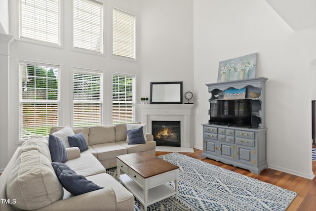 living room with high vaulted ceiling, healthy amount of natural light, a glass covered fireplace, wood finished floors, and baseboards