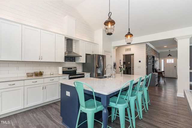 kitchen featuring stainless steel appliances, wall chimney exhaust hood, decorative columns, a sink, and high vaulted ceiling
