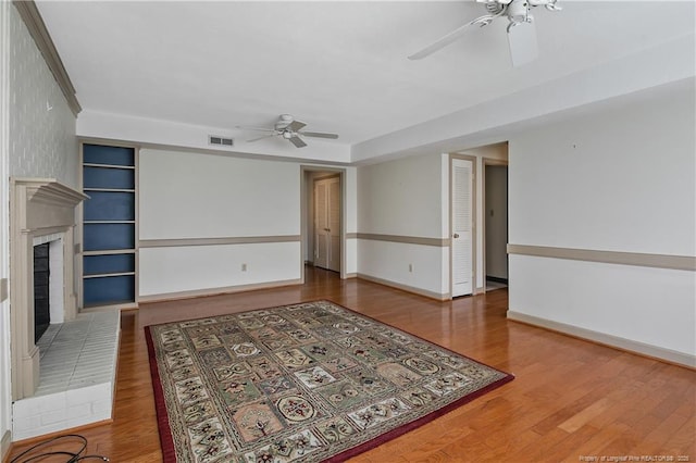 unfurnished living room with a brick fireplace, a ceiling fan, visible vents, and wood finished floors