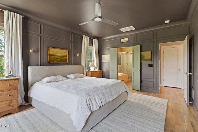 bedroom with a decorative wall, light wood-style flooring, ensuite bath, and a ceiling fan