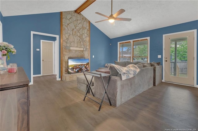 living area with a ceiling fan, baseboards, a stone fireplace, wood finished floors, and vaulted ceiling with beams