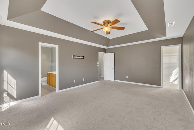 unfurnished bedroom with a raised ceiling, light colored carpet, baseboards, connected bathroom, and ceiling fan