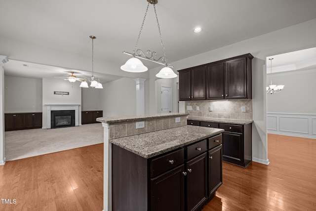 kitchen with a ceiling fan, wood finished floors, a center island, a glass covered fireplace, and light stone counters
