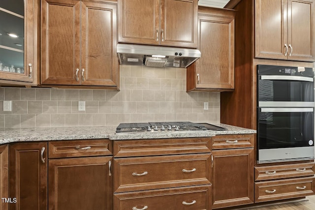 kitchen featuring under cabinet range hood, stainless steel gas cooktop, backsplash, glass insert cabinets, and light stone counters