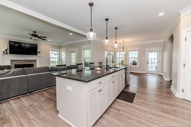 kitchen with light wood finished floors, ceiling fan, a sink, dark stone countertops, and a kitchen island with sink