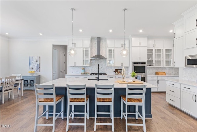 kitchen with wall chimney exhaust hood, stainless steel appliances, a kitchen island with sink, decorative backsplash, and white cabinetry