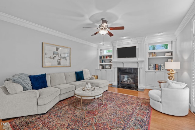 living room with light wood-type flooring, a ceiling fan, a tiled fireplace, and ornamental molding