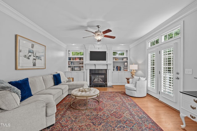 living room featuring light wood-style flooring, ceiling fan, a tile fireplace, ornamental molding, and built in shelves