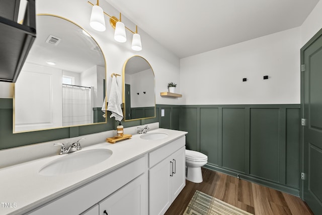 bathroom featuring wainscoting, wood finished floors, toilet, double vanity, and a decorative wall