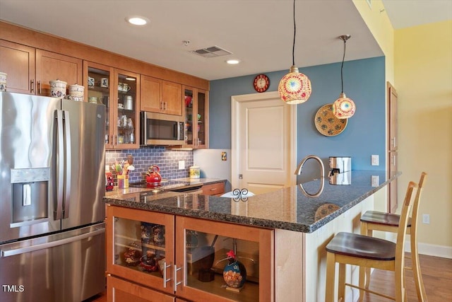 kitchen featuring stainless steel appliances, brown cabinetry, backsplash, glass insert cabinets, and dark stone counters
