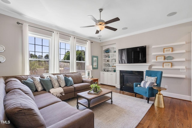 living area featuring crown molding, a ceiling fan, built in shelves, a glass covered fireplace, and wood finished floors