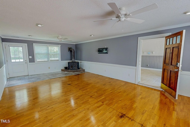 unfurnished living room with light wood-style flooring, a wood stove, a textured ceiling, ceiling fan, and crown molding