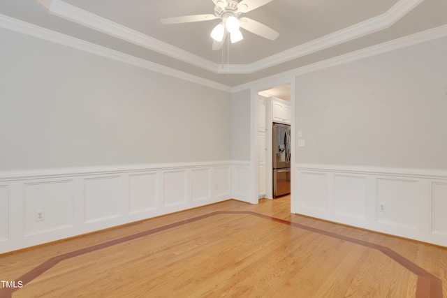 empty room with light wood-style flooring, ceiling fan, crown molding, wainscoting, and a raised ceiling