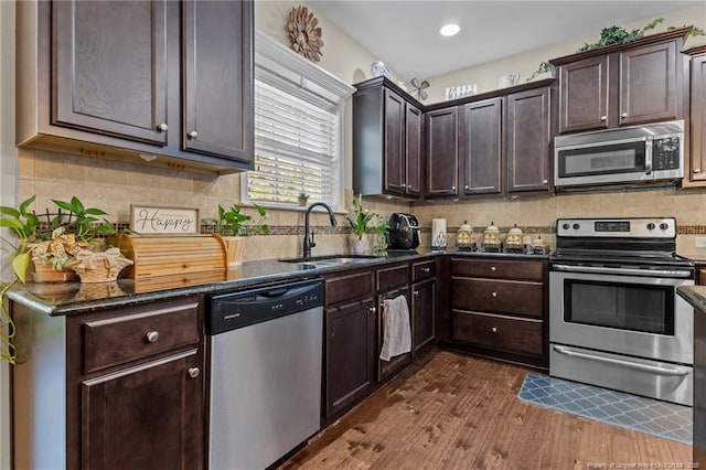 kitchen featuring stainless steel appliances, a sink, dark brown cabinetry, dark wood finished floors, and dark stone counters