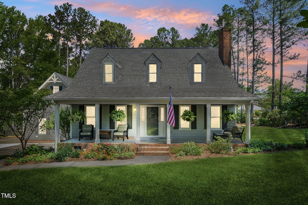 cape cod home with a porch, a lawn, a chimney, and roof with shingles