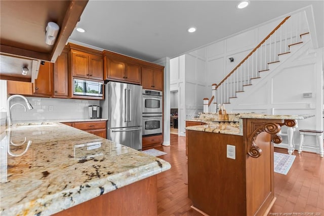 kitchen with a sink, brown cabinetry, a decorative wall, light stone countertops, and appliances with stainless steel finishes