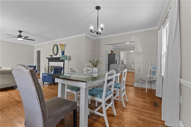dining room with light wood finished floors, a fireplace, ceiling fan with notable chandelier, and ornamental molding