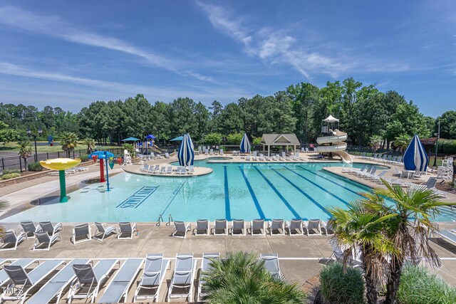 community pool featuring a water slide, a water play area, fence, a patio area, and view of wooded area
