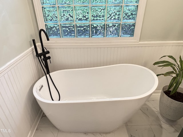 full bath featuring marble look tile flooring, a wainscoted wall, and a freestanding tub