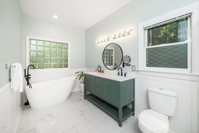 bathroom featuring wainscoting, marble look tiles, a freestanding tub, toilet, and double vanity