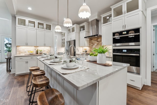 kitchen with wall chimney range hood, a center island with sink, white cabinetry, freestanding refrigerator, and dark wood-style flooring
