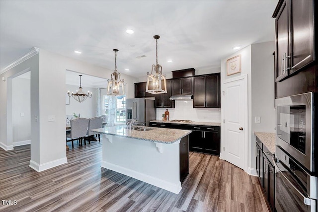 kitchen with a sink, stainless steel appliances, under cabinet range hood, wood finished floors, and a chandelier