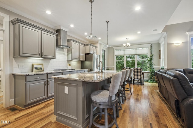 kitchen featuring gray cabinets, wall chimney exhaust hood, open floor plan, crown molding, and a kitchen island with sink