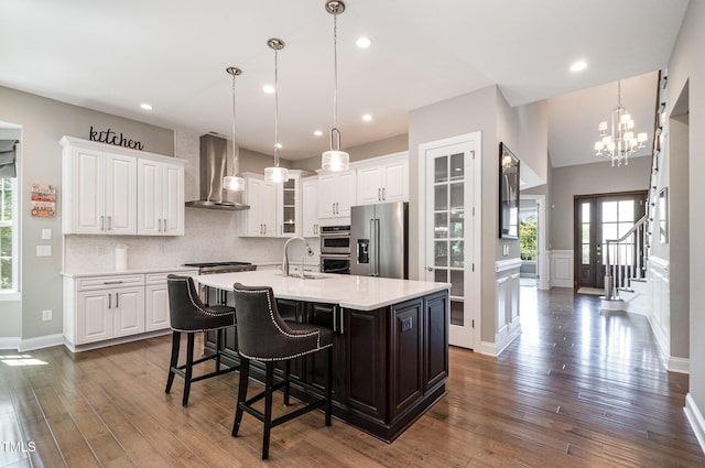 kitchen featuring stainless steel appliances, wall chimney exhaust hood, light countertops, an island with sink, and white cabinetry