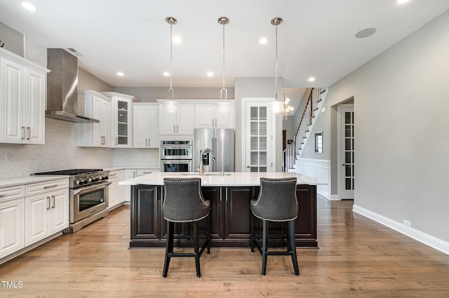 kitchen featuring stainless steel appliances, wall chimney range hood, a kitchen island with sink, light wood-style floors, and light countertops