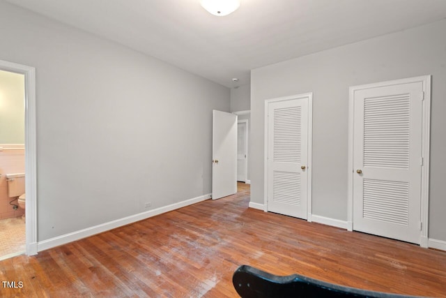 unfurnished bedroom with wood finished floors, two closets, baseboards, and ensuite bath