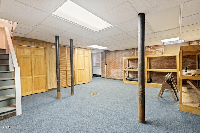 finished basement with brick wall, carpet floors, a drop ceiling, and stairs