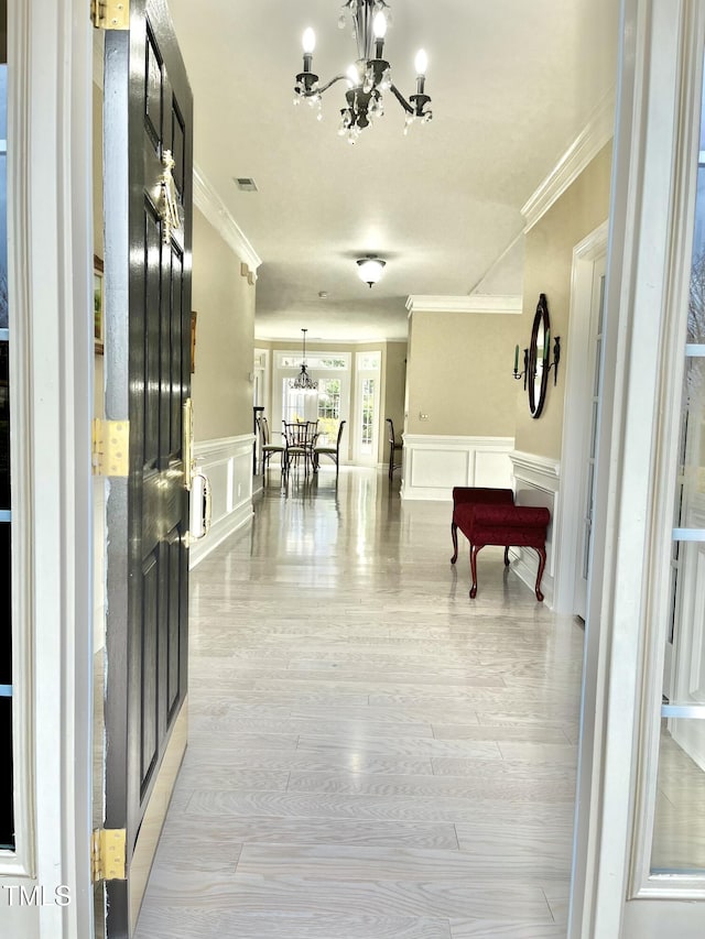 corridor featuring a chandelier, crown molding, a decorative wall, wainscoting, and light wood-style floors