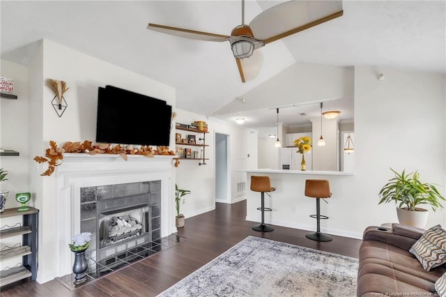 living room with a ceiling fan, vaulted ceiling, dark wood-type flooring, a tile fireplace, and baseboards